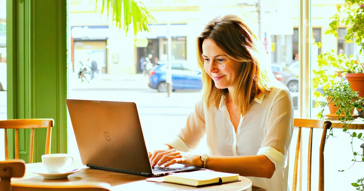 Person working on laptop in a sunlit cafe with large windows and plants
