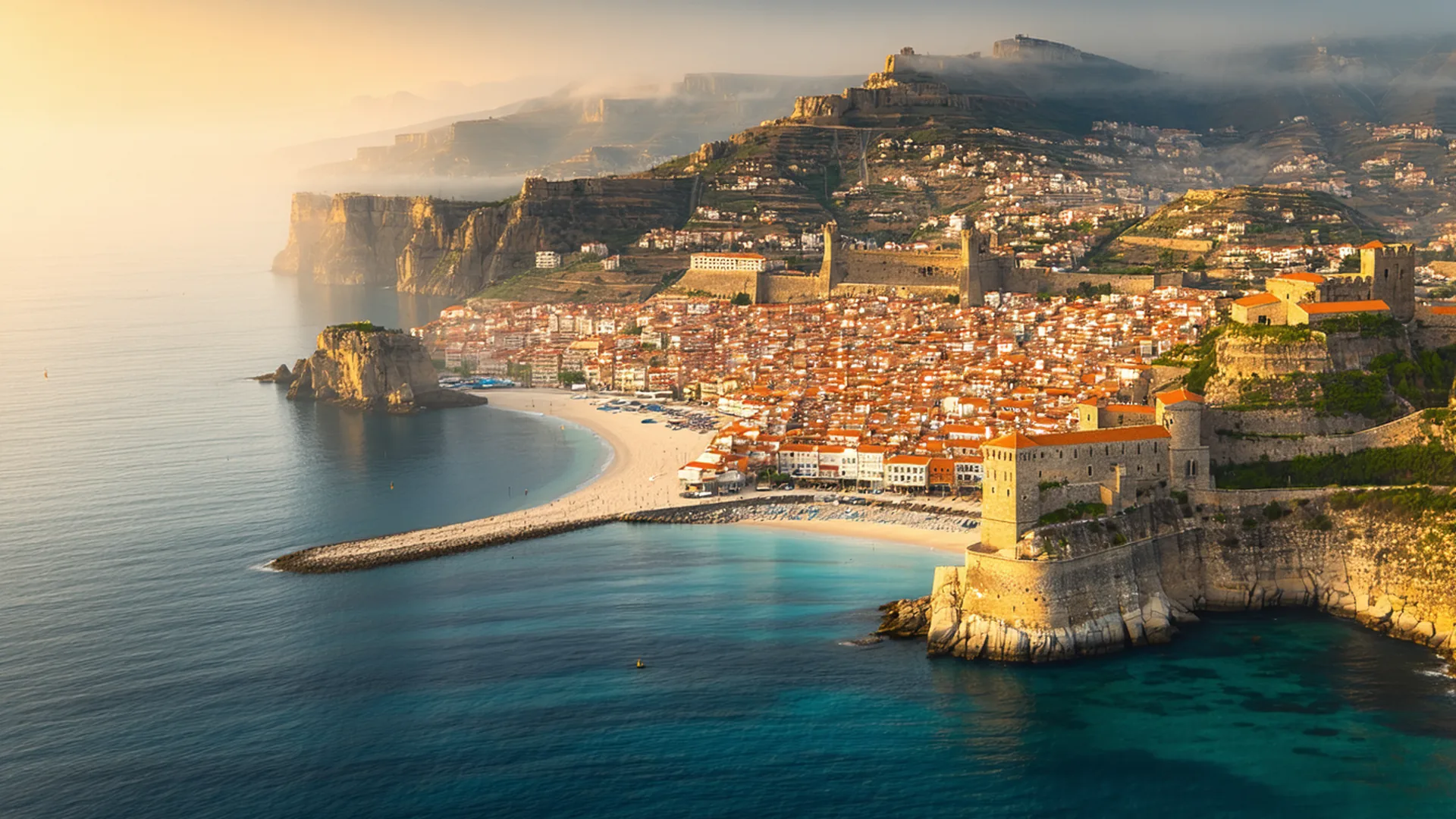 Golden-hour view of terracotta rooftops and coastline from a hilltop viewpoint, warm amber light